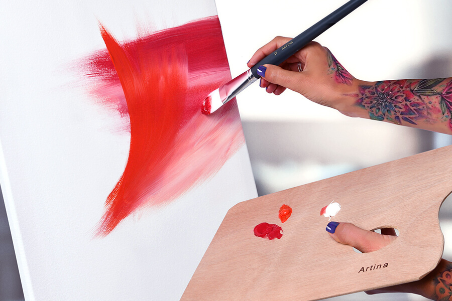 Frau malt mit roter Acrylfarbe auf weiße Leinwand und hält eine Holzpalette in der Hand