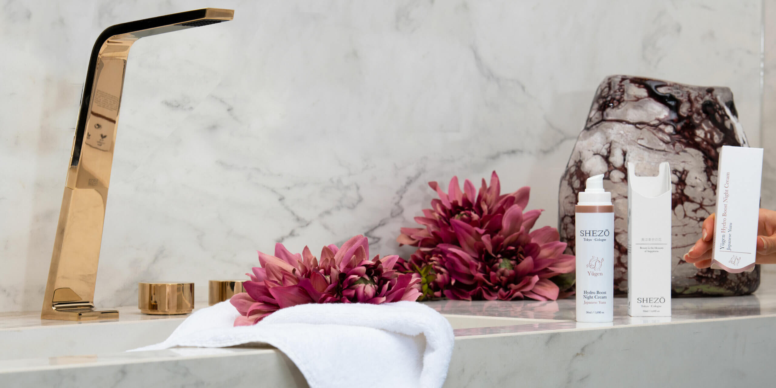 Waschbecken mit goldenem Wasserhahn, an dem Shezo Produkte stehen und lilane Blumen liegen.